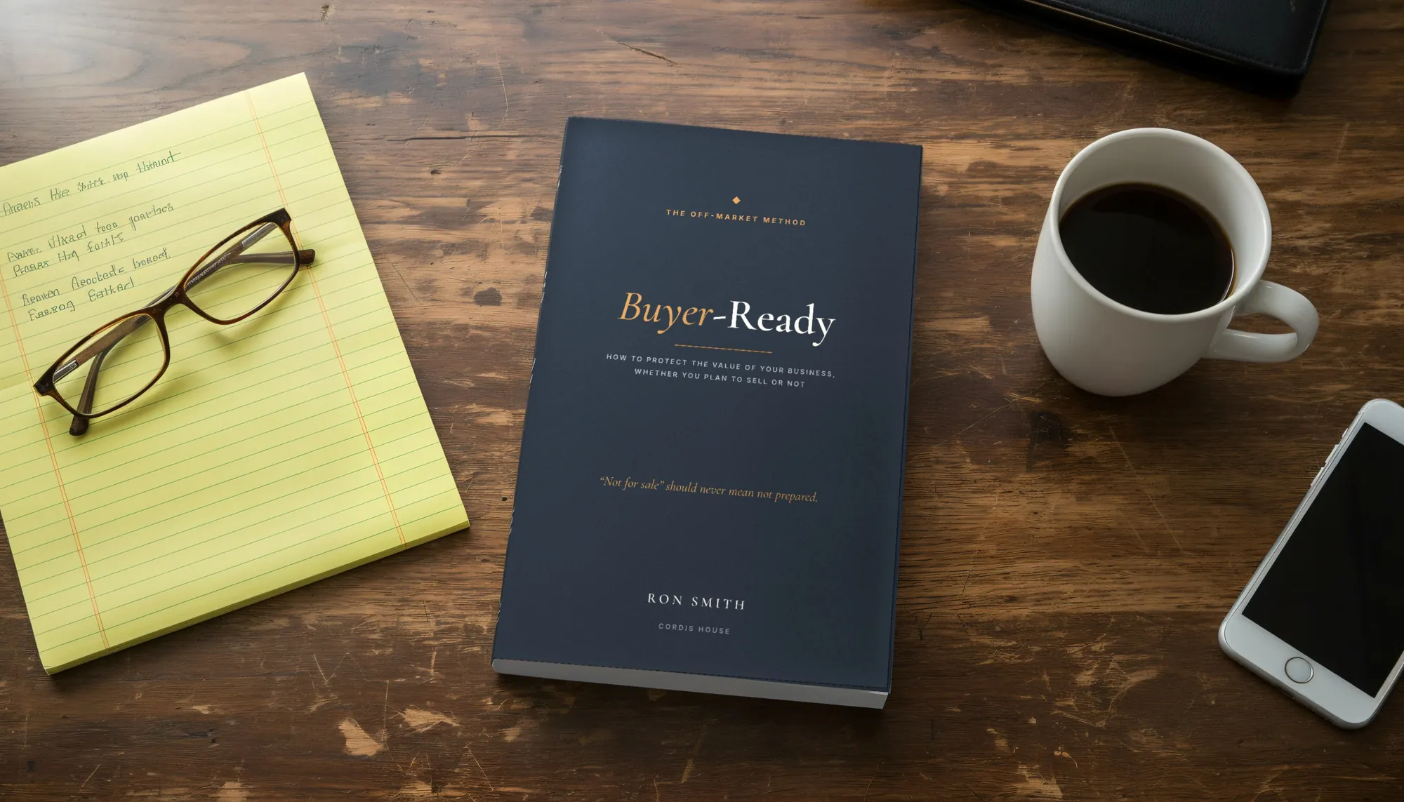 A paperback of Buyer-Ready on a wooden desk beside a yellow legal pad, reading glasses, a cup of coffee and a phone